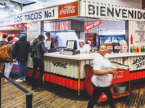 The Los Tacos No. 1 counter inside Chelsea Market, with the hand-painted 'Los Tacos No.1' and 'Bienvenidos' signs, aguas frescas jars, and customers ordering