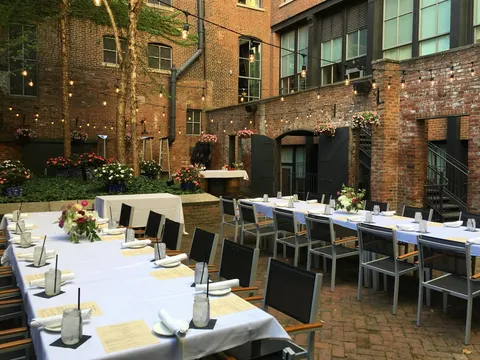 Outdoor brick courtyard patio at Mussel & Burger Bar Downtown in Louisville, set with long white-linen tables, black chairs, mason-jar water glasses and flowers under strung café lights