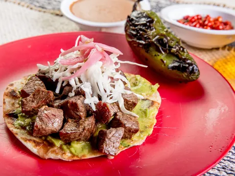 Mesquite-grilled carne asada taco on a red plate with guacamole, pickled onion and cabbage, a charred poblano and refried beans, at Casa Asada Taqueria y Cerveceria in Tucson