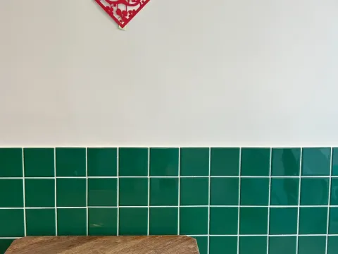 Interior corner at Ho Foods in New York — a wooden table against a green-tiled wall with a red paper-cut good-fortune decoration and a teal stool