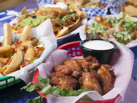 A spread of Cilantro's Mexican Restaurant dishes on a blue table — a basket of fried chicken wings with ranch, bacon-and-cheese loaded fries, guacamole nachos and street tacos