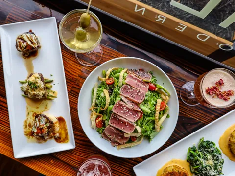 Overhead spread of dishes at ML Steak Modern Chophouse, including a seared tuna salad, filet medallions, a crab cake and cocktails on a wood table
