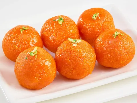 Six bright orange motichoor laddoo, ghee-fried gram-flour sweet balls topped with slivered pistachio, on a white plate, from Mithaas in Edison, New Jersey