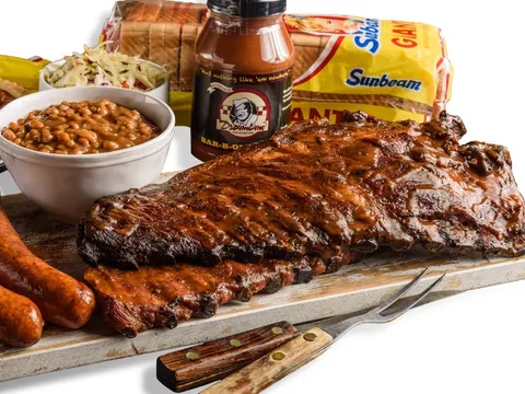 Dreamland Bar-B-Que full slab of ribs on a cutting board with two smoked sausage links, a bowl of baked beans, coleslaw, a pecan pie, a bottle of Dreamland BBQ sauce and Sunbeam bread