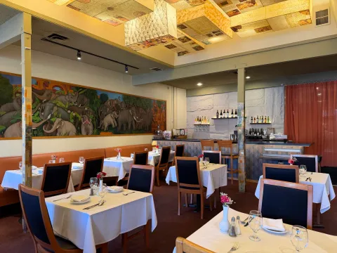 Ajanta's dining room in Berkeley with white-clothed tables, a long elephant mural, and a wine bar under a gilded panel ceiling