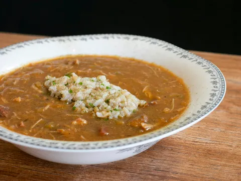 A bowl of gumbo topped with a scoop of white rice and chopped chives, served in a patterned-rim white bowl on a wooden table at Virtue in Chicago