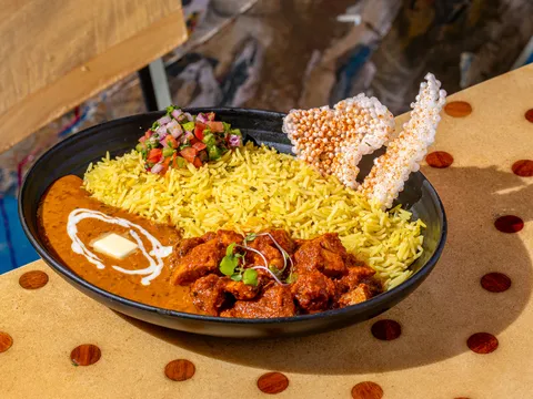 A Curry Up Now bowl of butter chicken makhni with turmeric rice, pico kachumber and puffed sabudana papad in a black bowl, brand food photograph