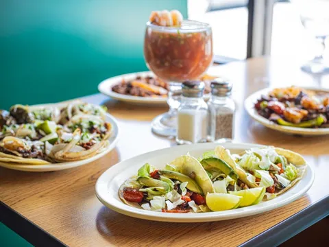 Plates of tacos with avocado and lettuce alongside a shrimp cocktail on a table at Taqueria Los Compadres