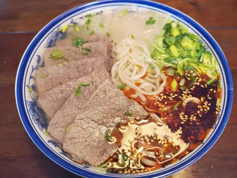 A bowl of Kung Fu Noodle's Classic Lanzhou beef noodle soup with sliced beef, hand-pulled noodles, cilantro, garlic greens and chili oil in a blue-and-white porcelain bowl