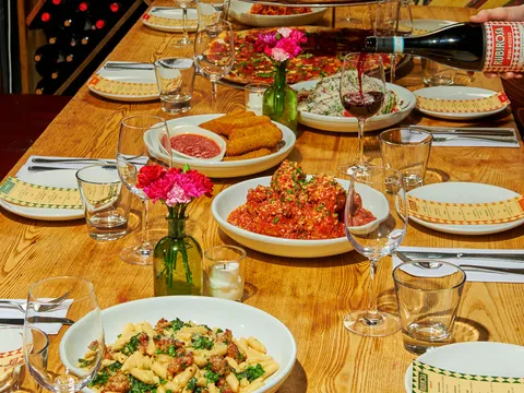 Family-style spread at Rubirosa with the signature tie-dye pizza, cavatelli, meatballs and a bottle of Rubi Rosso wine being poured on a wooden table