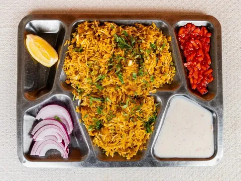 A biryani thali on a steel compartment tray at Punjabi Dhaba in Cambridge, with saffron-hued rice, sliced red onion, chopped tomato and raita